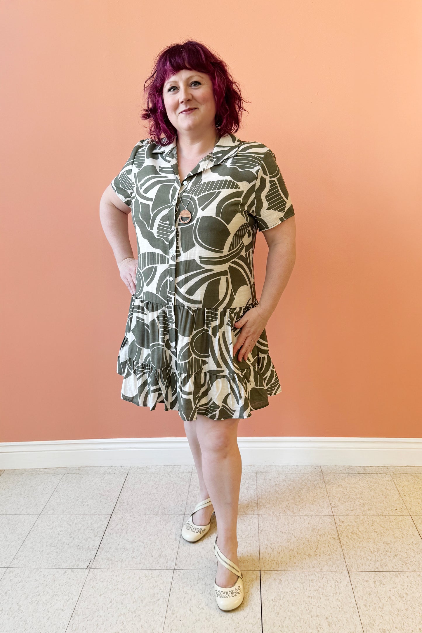 Allana is wearing the Olympia Dress by Compli K in a Green/Cream geometric print, a short-sleeved dress with a blouse collar, button front, short tiered skirt with ruffles, and a slip dress under-layer. She is standing in front of a coral wall.