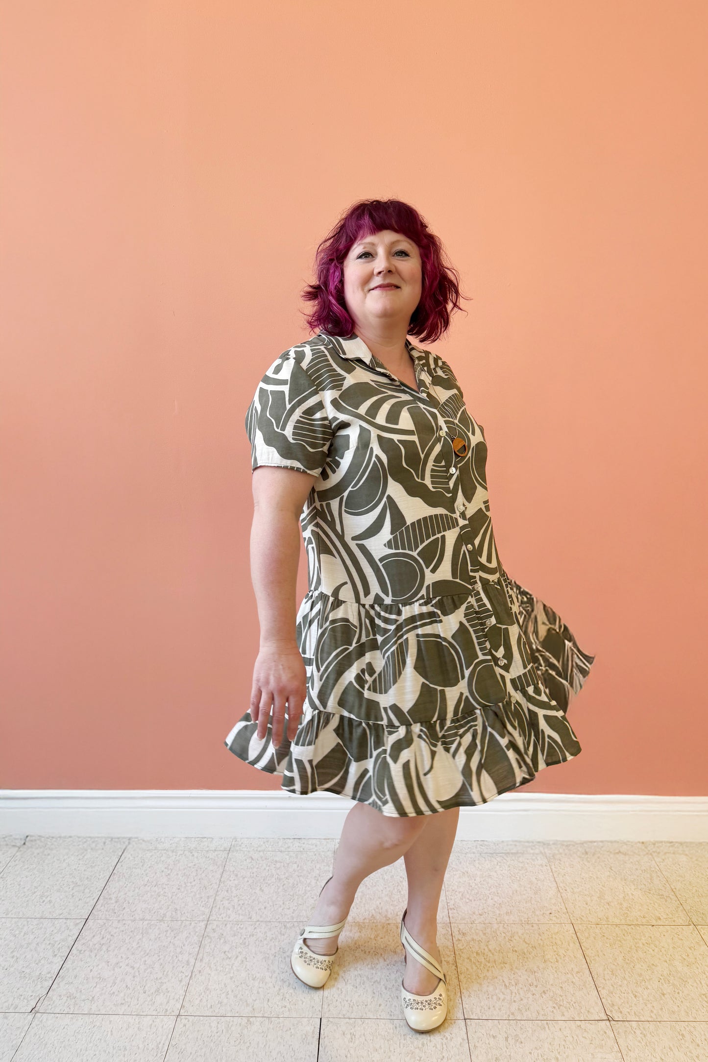 Allana is wearing the Olympia Dress by Compli K in a Green/Cream geometric print, a short-sleeved dress with a blouse collar, button front, short tiered skirt with ruffles, and a slip dress under-layer. She is standing in front of a coral wall.