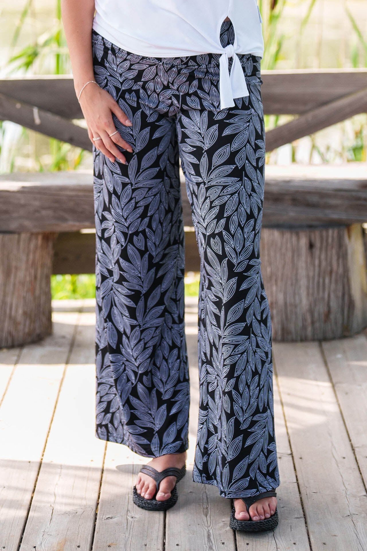 A waist-down view of a woman wearing the Rien ne se Perd Rumba Palazzo Pants in Black and White Foliage with a white top, standing inside a wooden gazebo.
