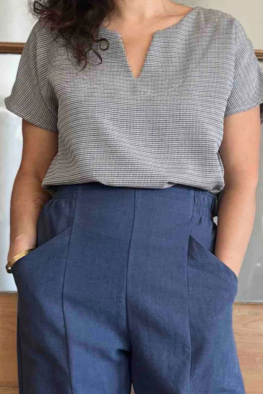 Neck down view of a woman wearing the Lucca Top in Blue Check print by Kazak, a short sleeved top with a notched V-neckline and a pleated yoke at the back. She is wearing it with the Sienna Pants in Lapis, and is standing in front of a wooden door.