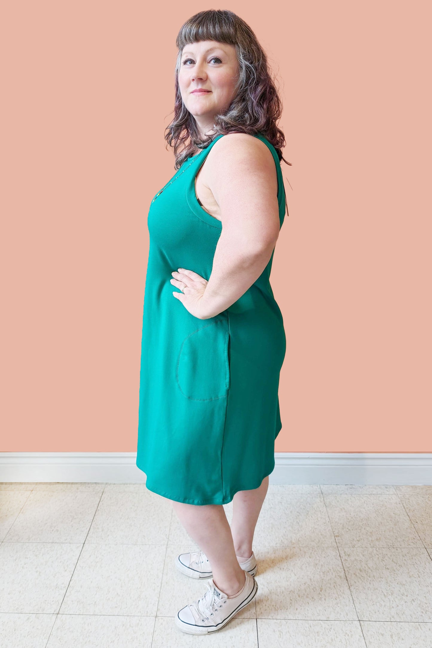 Side view of Allana wearing the Cheri Dress by Pure Essence in Kelly Green, standing in front of a coral wall