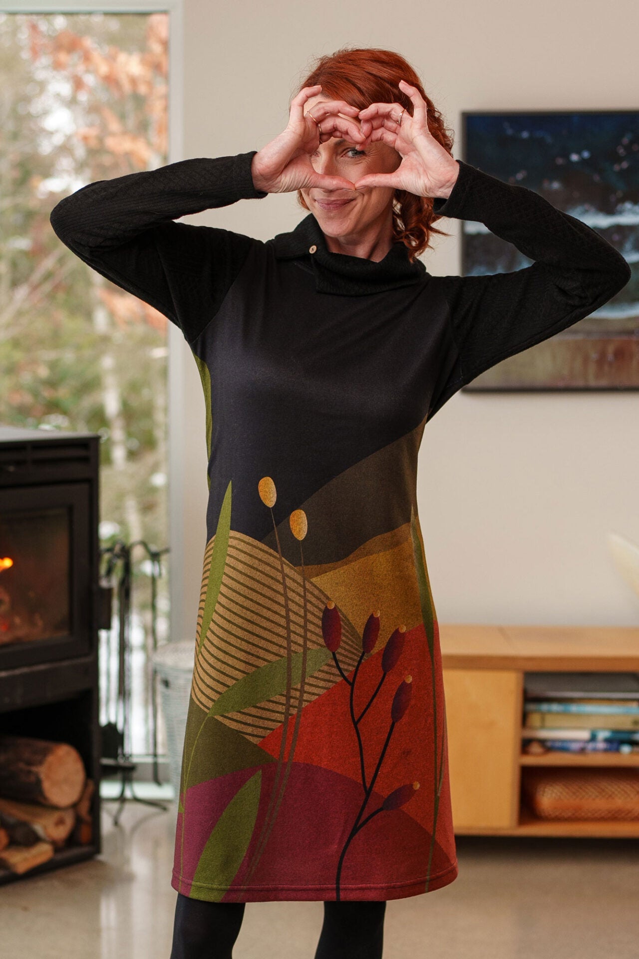 Waist up view of a woman wearing the Cornouiller Dress by Rien ne se Perd in Paysage print, featuring textured black fabric on the upper bodice and long sleeves and a landscape print on the body. It has a cowl neck with a slit and coconut button detail. She is standing in a room with a fireplace.