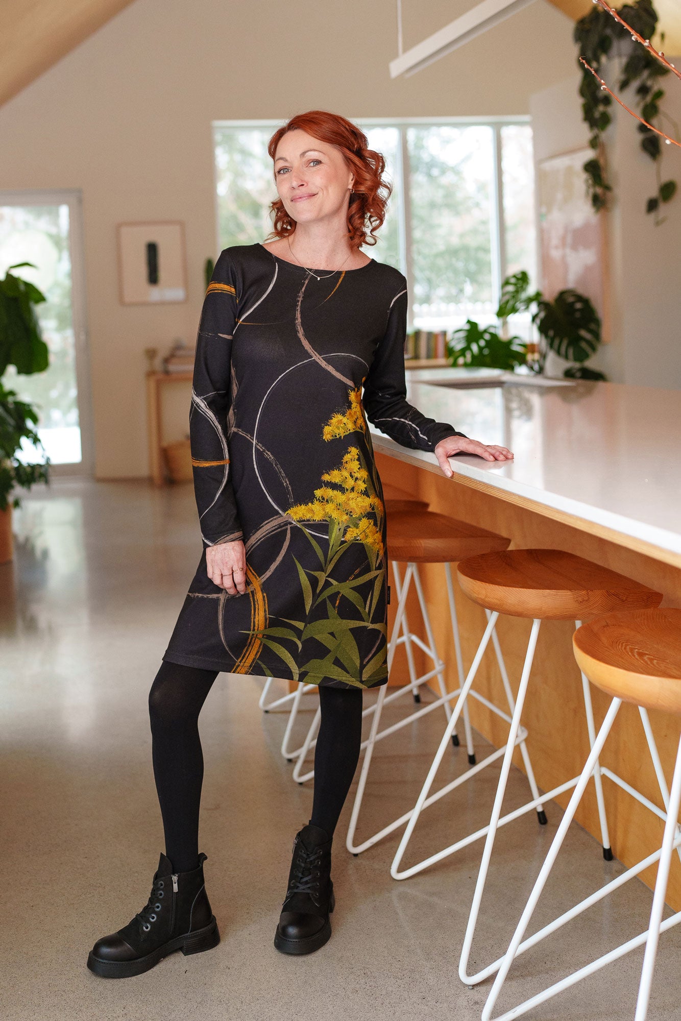 A woman wearing the Flore Dress by Rien ne se Perd in Bordure, a semi-fitted black dress with a flora inspired print, long sleeves, a round neck, and hem that hits above the knees. She is standing in a kitchen.
