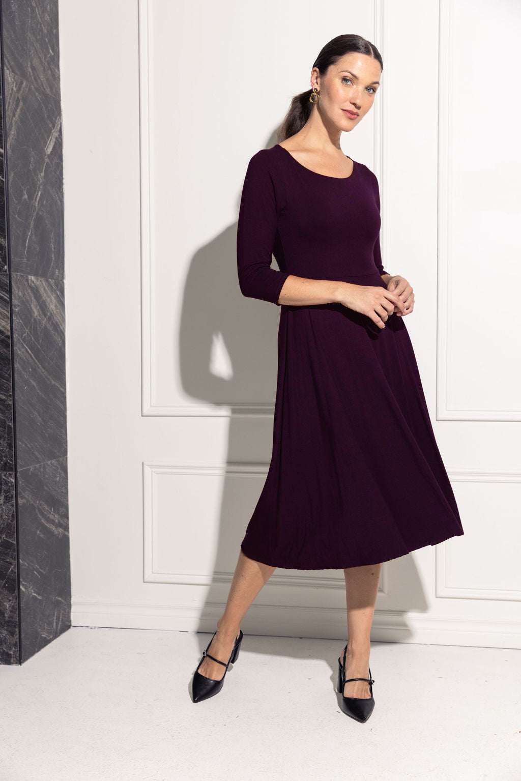 A woman wearing the Diamond Dress by Cherry Bobin in Purple, featuring a boat neck, 3/4 sleeves, a fit and flare shape, and a tea-length skirt in bamboo rayon. She is standing. in front of a white wall.