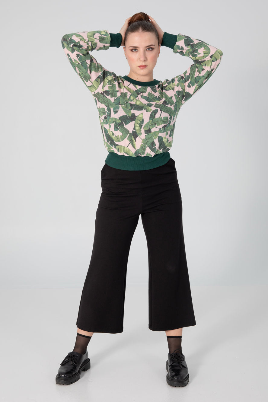A woman wearing the Wanda Sweater by Slak in Pink and Green Tropical Leaves, featuring long sleeves, a round neck, a semi-fitted shape, and contrasting hem, cuffs and collar. She is wearing it with black pants and standing in front of a grey background.