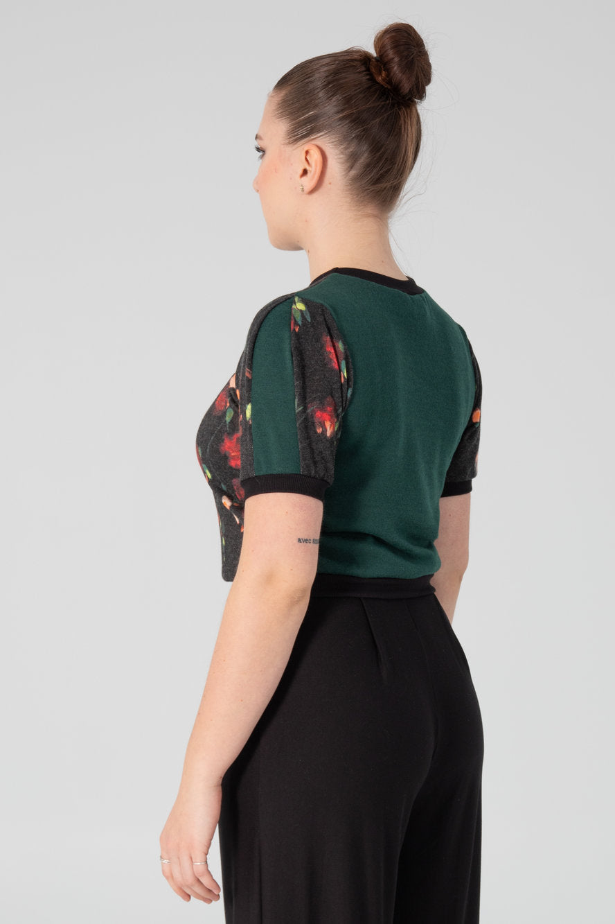A woman wearing the Darling T-Shirt by Slak in Floral, a short sleeved, semi-fitted sweater with contrasting fabric at the cuffs, collar and hem, inserts on the sleeves, and a solid back. She is wearing it with black pants and standing in front of a white background.
