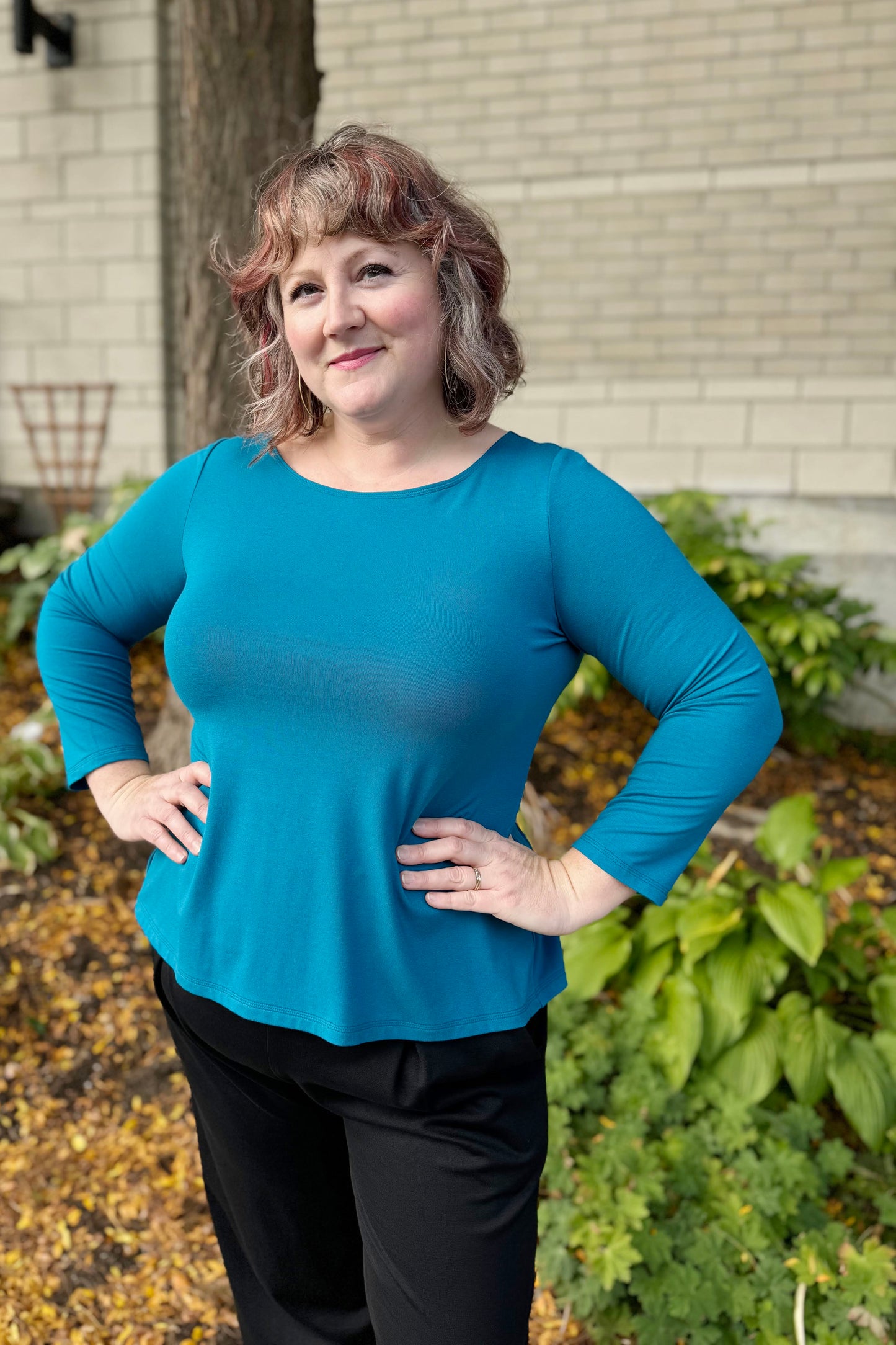 A woman wearing the Studio D Fela top in Teal, featuring 3/4 sleeves and a split hem. She is wearing the Wilfred Pants by Melow in Black and standing in front of a garden.