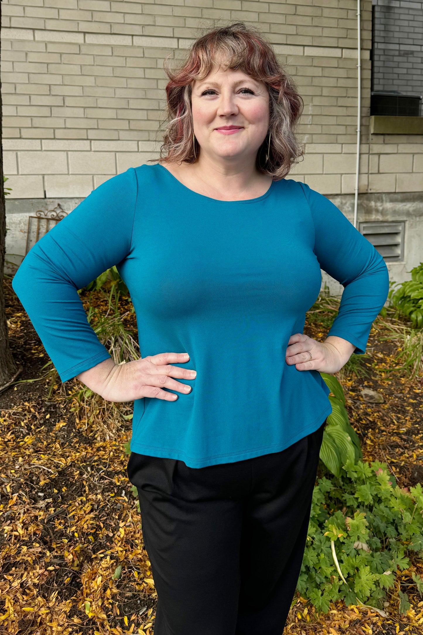 A woman wearing the Studio D Fela top in Teal, featuring 3/4 sleeves and a split hem. She is wearing the Wilfred Pants by Melow in Black and standing in front of a garden.