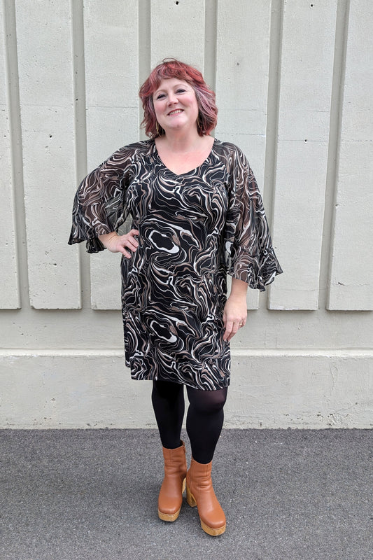 A woman wearing the Nyla Dress by Compli K in Black/Beige Swirl, with a V-neck, fly-away chiffon sleeves with a slit detail, sheath shape, and above the knee hem. She is standing outside in front of a white wall.