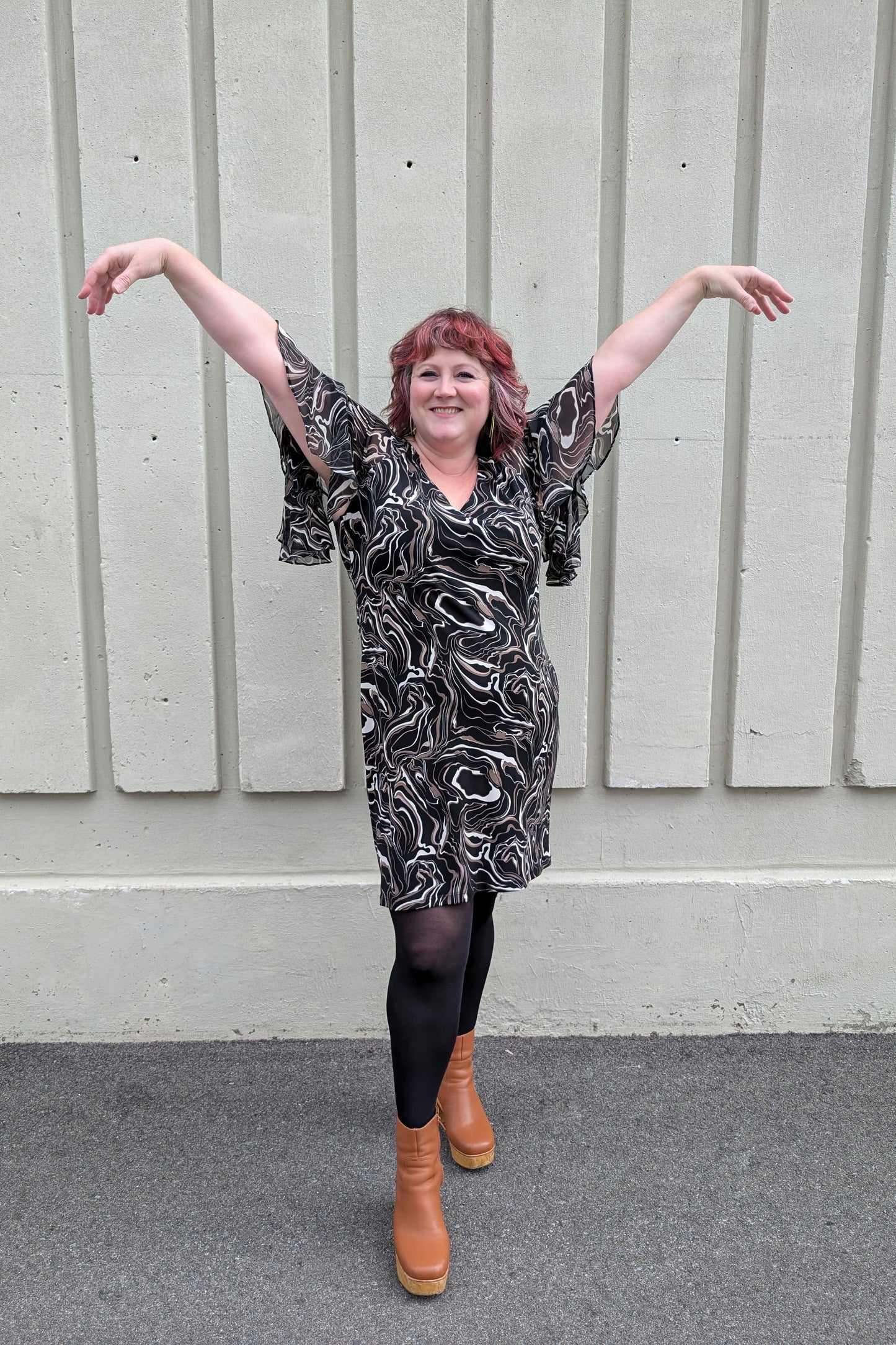 A woman wearing the Nyla Dress by Compli K in Black/Beige Swirl, with a V-neck, fly-away chiffon sleeves with a slit detail, sheath shape, and above the knee hem. She is standing outside in front of a white wall.