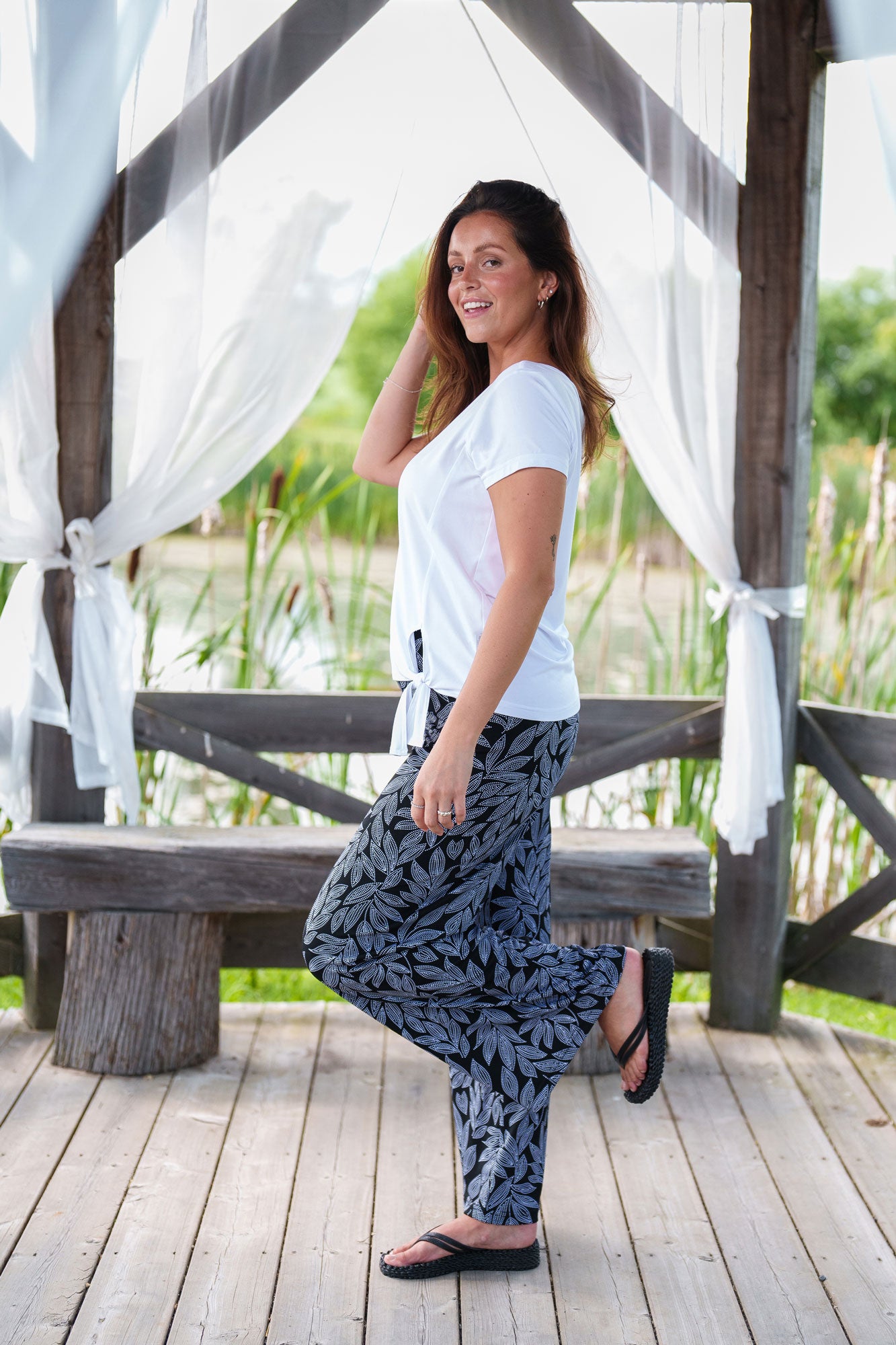 Side view of a woman wearing Rumba Palazzo Pants in Black and
White Foliage and a white top standing outside in a wooden gazebo.