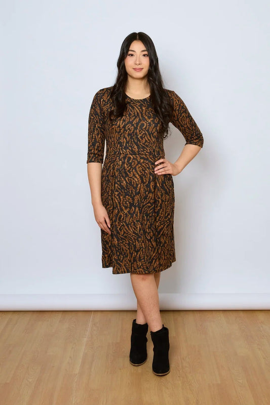 A woman wearing the Meadow Dress by Tangente in Rust Print, with a round neck, 3/4 sleeves, princess seams, and a pleated knee-length skirt. She is standing against a white wall.