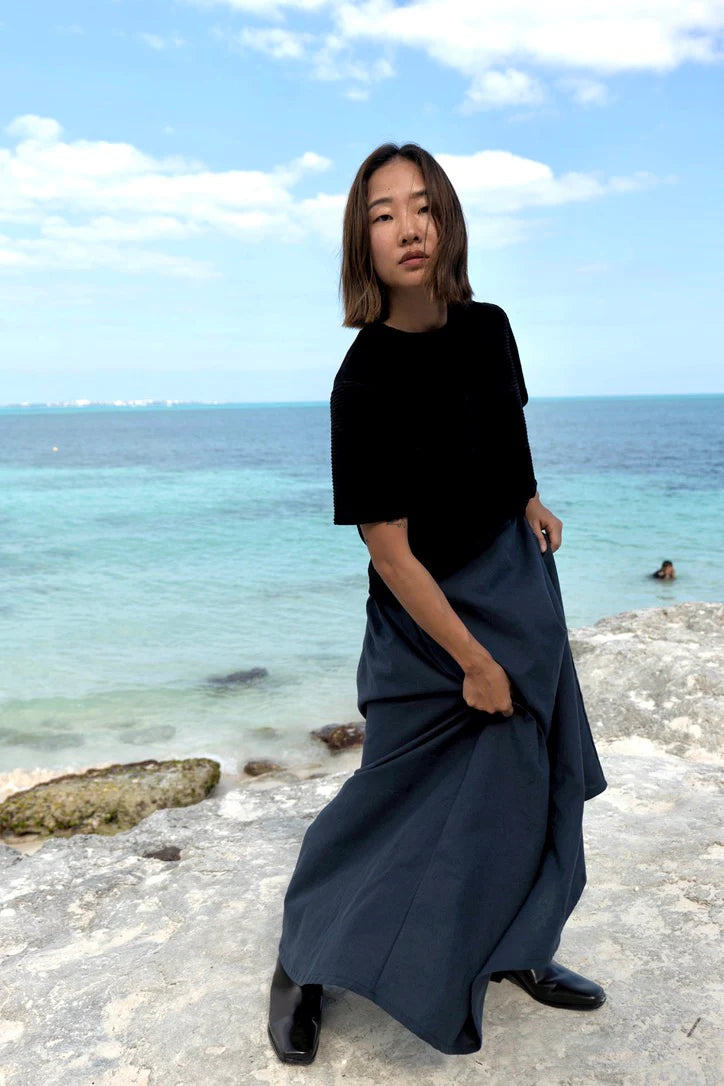 A woman wearing the Tofino T-Shirt by MAS in Pepper, a short sleeved top in ribbed velvet. She is wearing it with a blue skirt and standing on the beach.