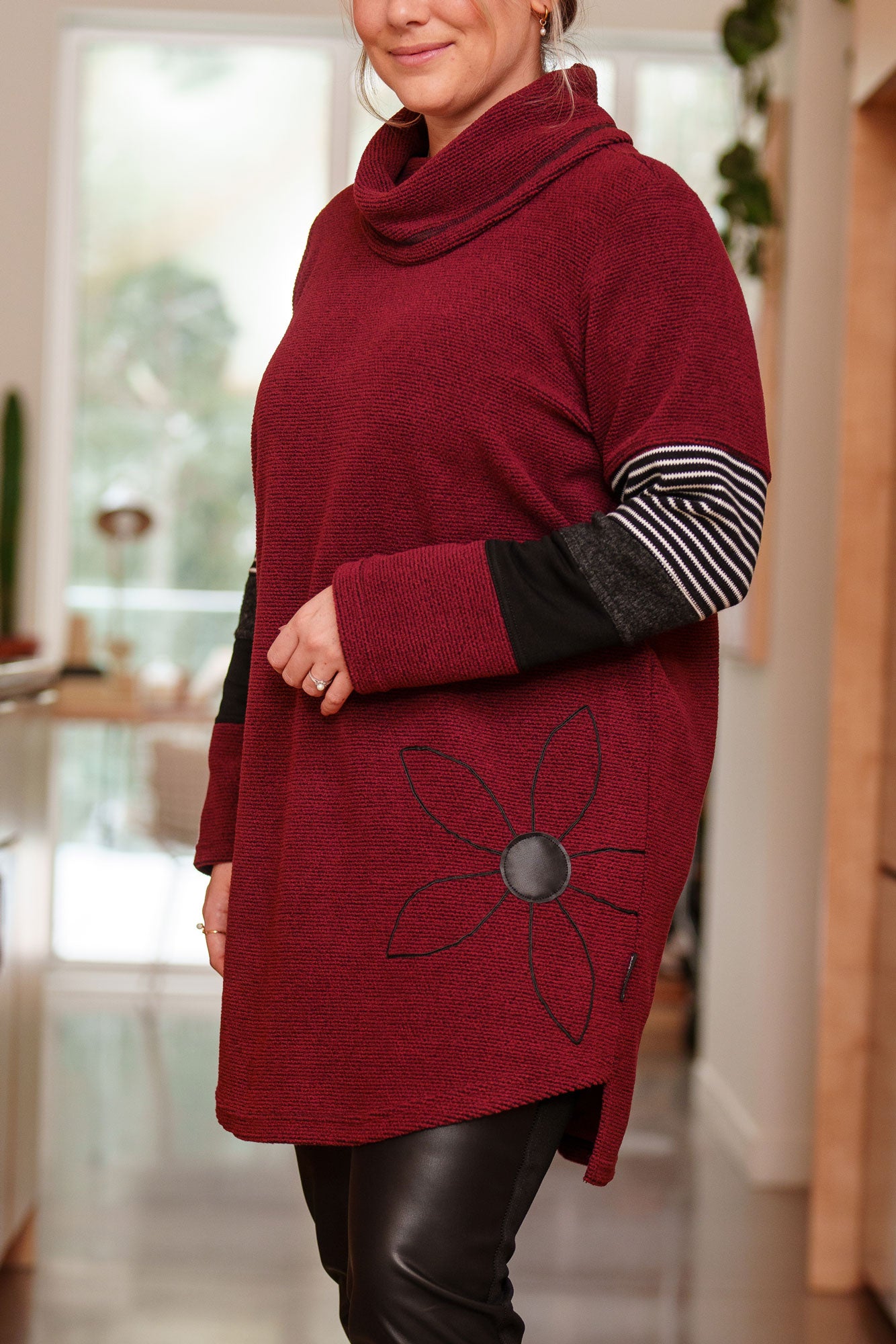 Side view of a woman wearing the Églantier Tunic by Rien ne se Perd in textured Bordeaux fabric with striped and black colour-blocking on the long sleeves. It has a large turtleneck, a faux leather floral decal, and a side pocket. She is wearing it with black leggings and standing in a kitchen.