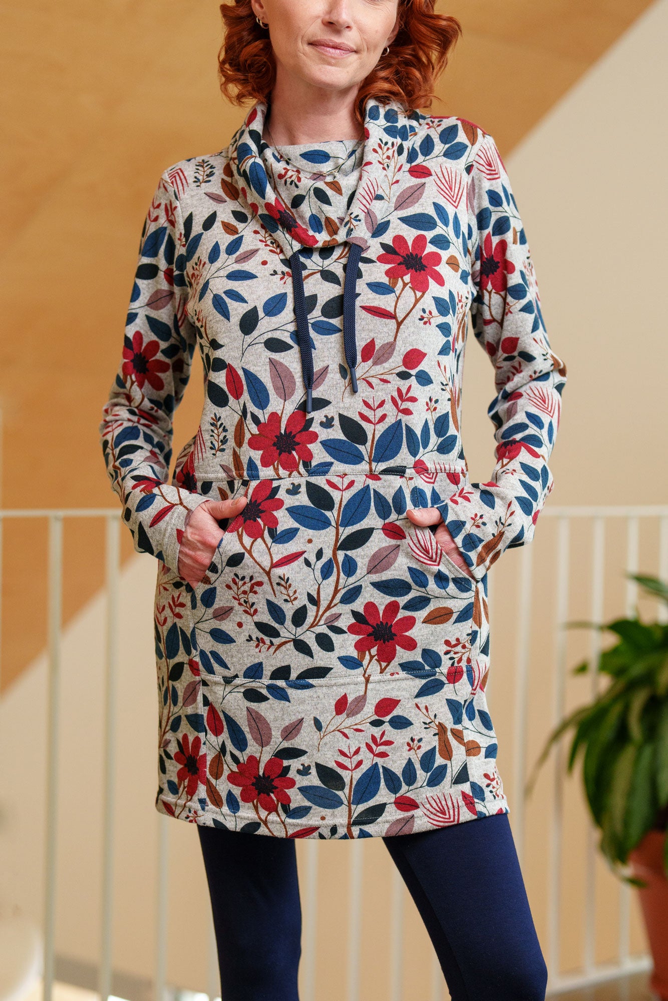 Torso view of a woman wearing the Genevrier Tunic by Rien ne se Perd in Floral, featuring a turtleneck with a drawstring, a kangaroo pocket, long sleeves, and a straight cut. She is wearing it with navy leggings and standing indoors with a plant behind her.