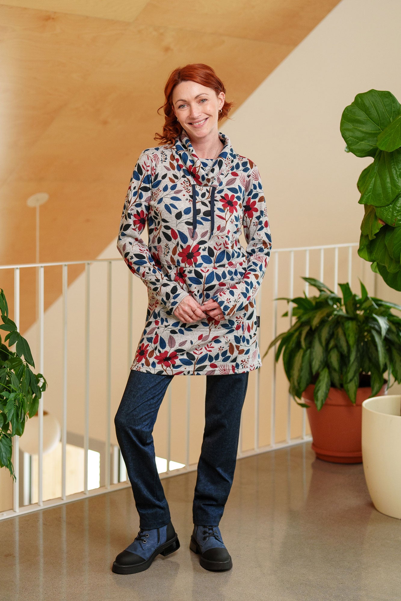 A woman wearing the Genevrier Tunic by Rien ne se Perd in Floral, featuring a turtleneck with a drawstring, a kangaroo pocket, long sleeves, and a straight cut. She is wearing it with navy leggings and standing indoors with plants behind her.