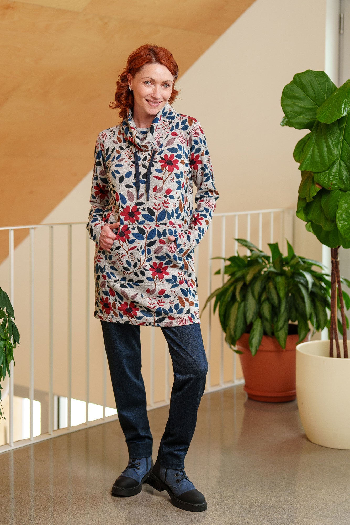 A woman wearing the Genevrier Tunic by Rien ne se Perd in Floral, featuring a turtleneck with a drawstring, a kangaroo pocket, long sleeves, and a straight cut. She is wearing it with jeans and standing indoors with plants behind her.