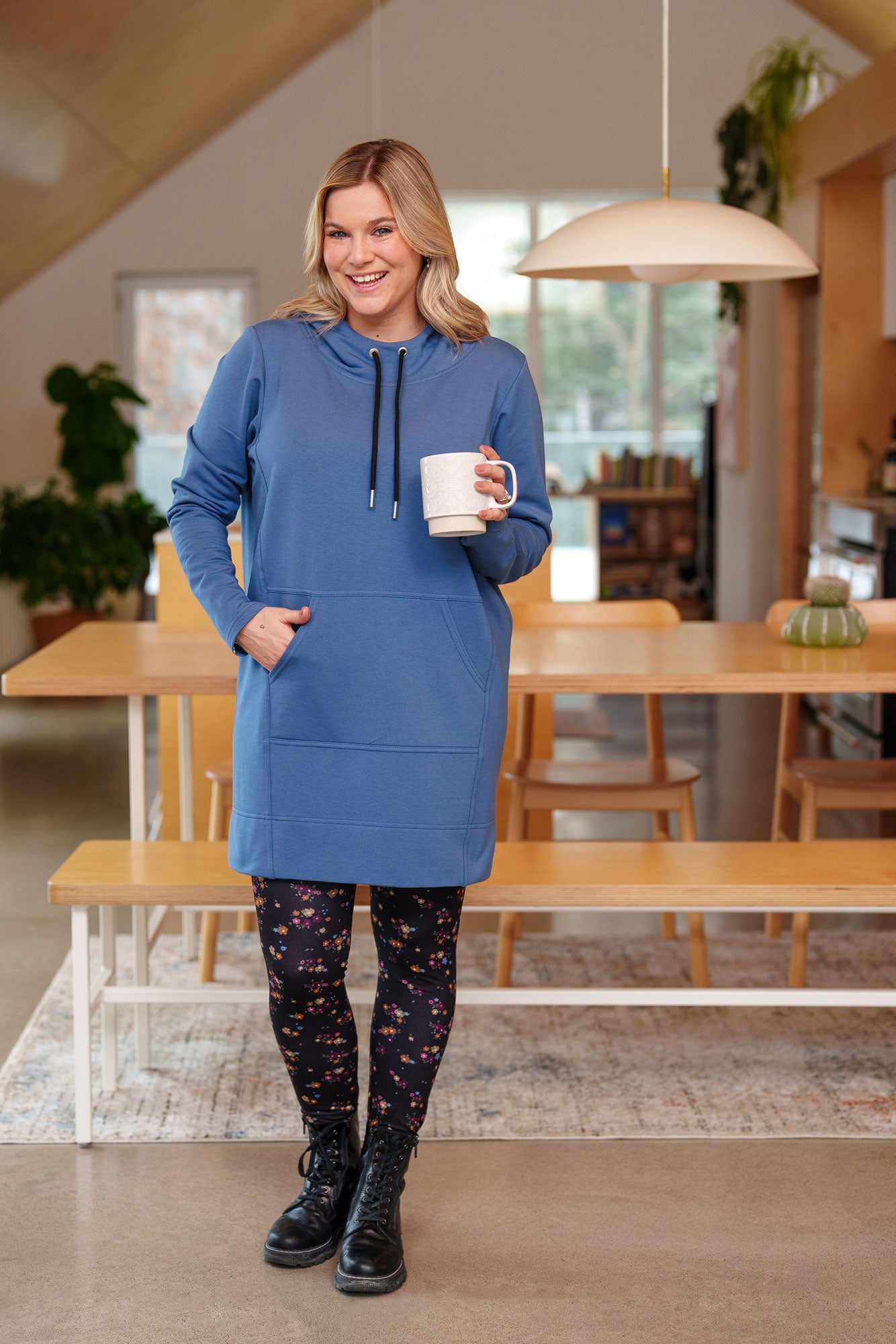 Torso view of a woman wearing the Toundra Tunic by Rien ne se Perd in Blue, featuring a large hood with drawstrings, a kangaroo pocket, and a relaxed fit. She is wearing it with the Laricin leggings and is standing in a kitchen holding a coffee mug.
