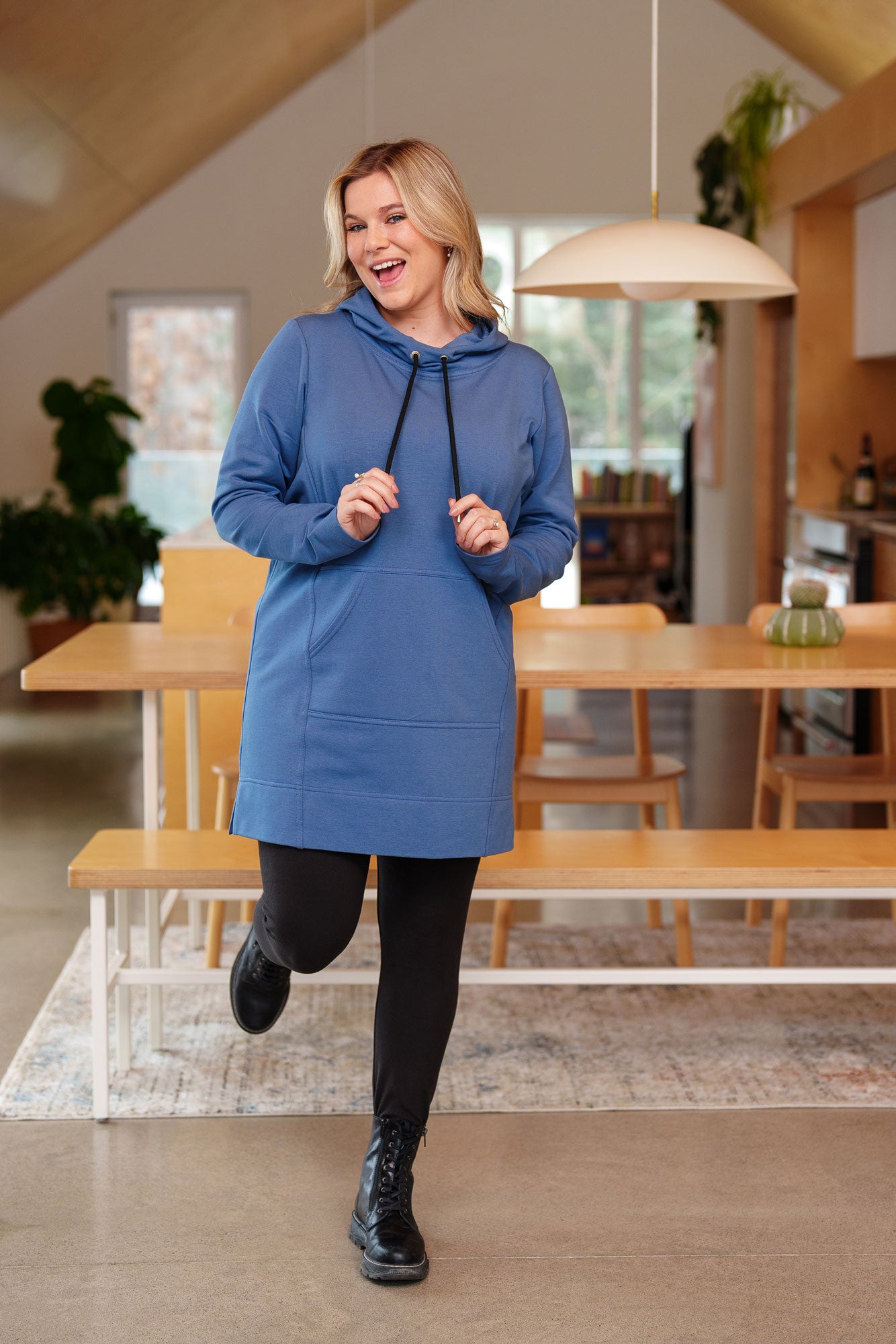 A woman wearing the Toundra Tunic by Rien ne se Perd in Blue, featuring a large hood with drawstrings, a kangaroo pocket, and a relaxed fit. She is wearing it with black leggings and is standing in a kitchen.