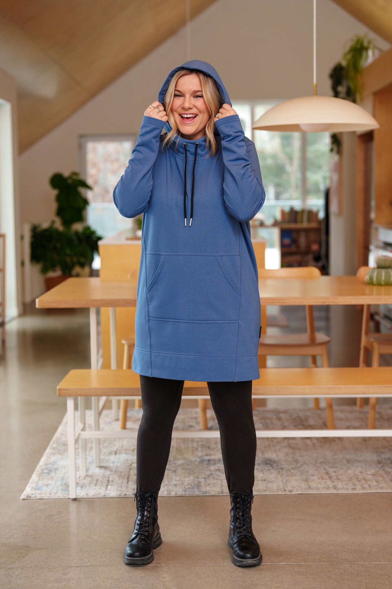 A woman wearing the Toundra Tunic by Rien ne se Perd in Blue, featuring a large hood with drawstrings, a kangaroo pocket, and a relaxed fit. She is wearing it with black leggings and is standing in a kitchen.