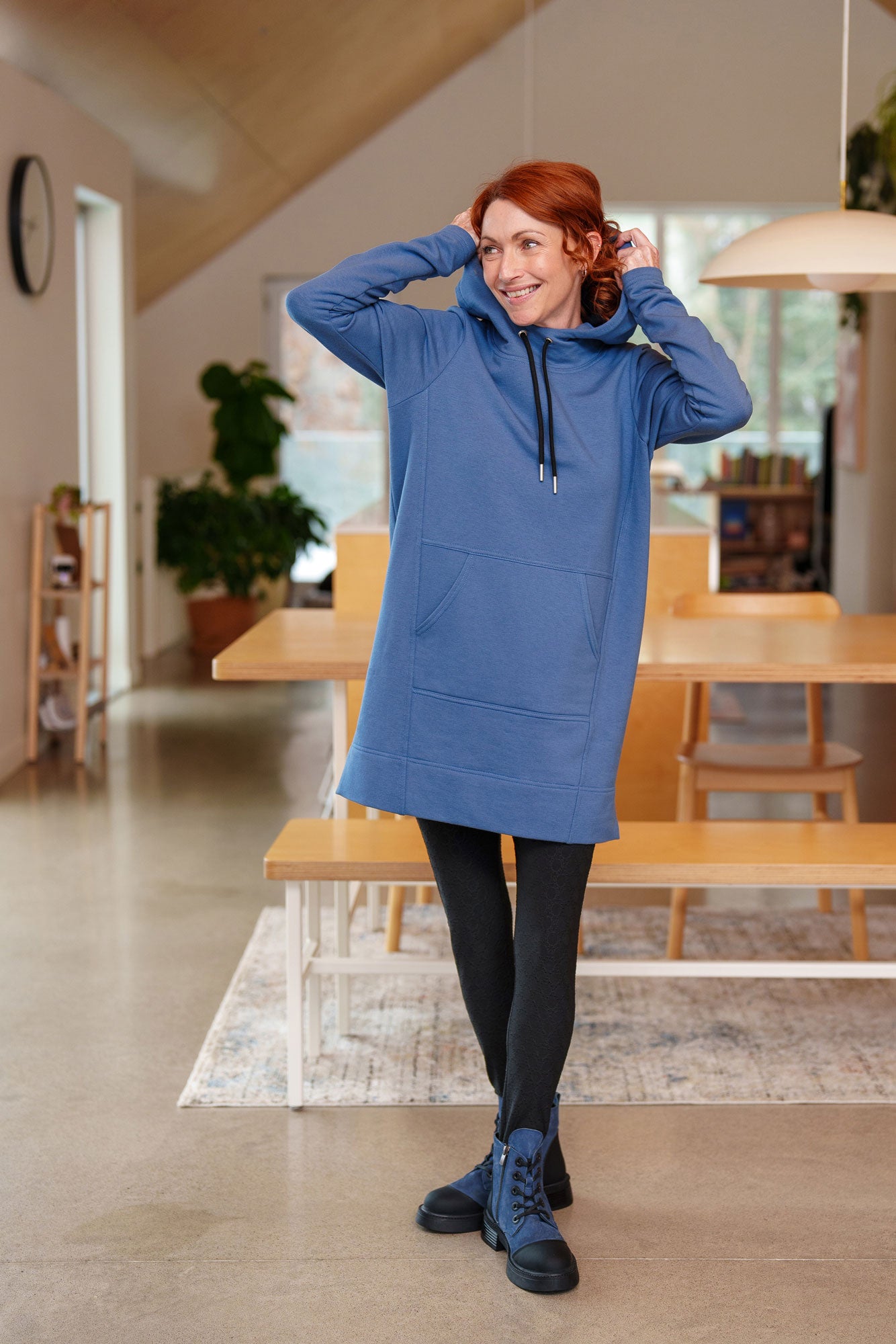 A woman wearing the Toundra Tunic by Rien ne se Perd in Blue, featuring a large hood with drawstrings, a kangaroo pocket, and a relaxed fit. She is wearing it with black leggings and is standing in a kitchen.
