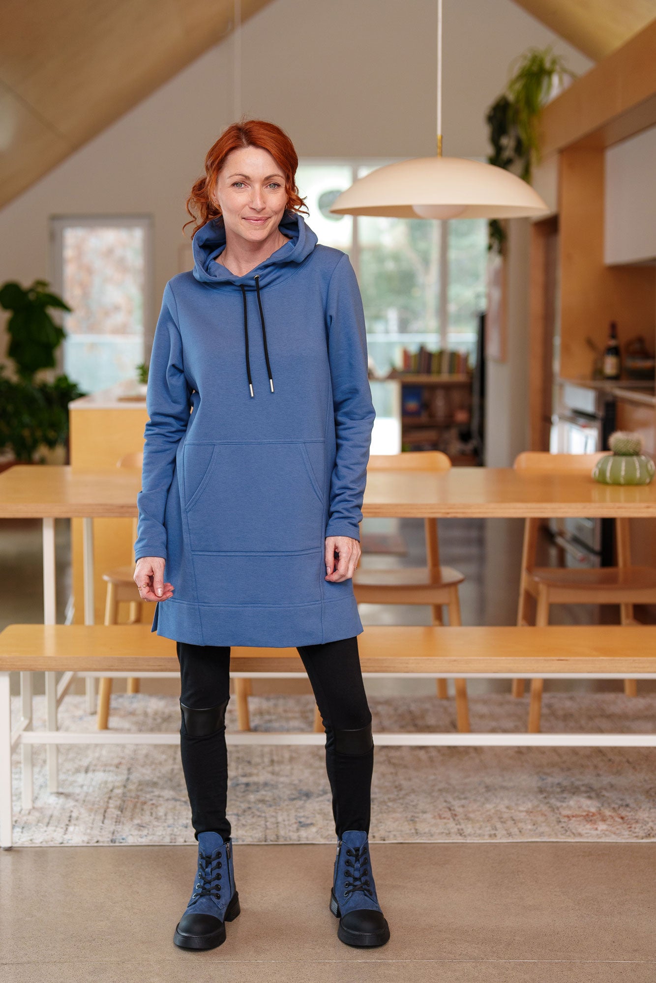 A woman wearing the Toundra Tunic by Rien ne se Perd in Blue, featuring a large hood with drawstrings, a kangaroo pocket, and a relaxed fit. She is wearing it with black leggings and is standing in a kitchen.