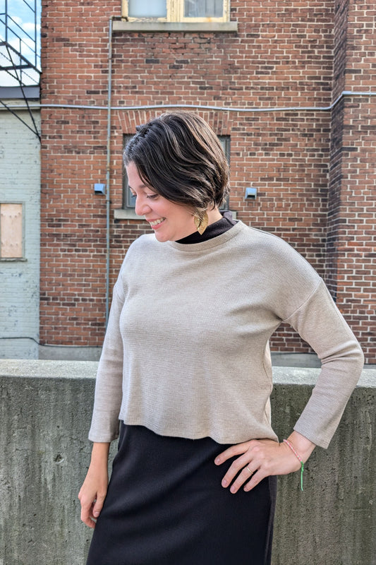 A woman wearing the Nerissa Knit Top by Compli K in Beige, with a round neck, long sleeves, slightly loose fit, and hi-low hemline, over the Natasha Dress in Black. She is standing outside in front of a brick wall.