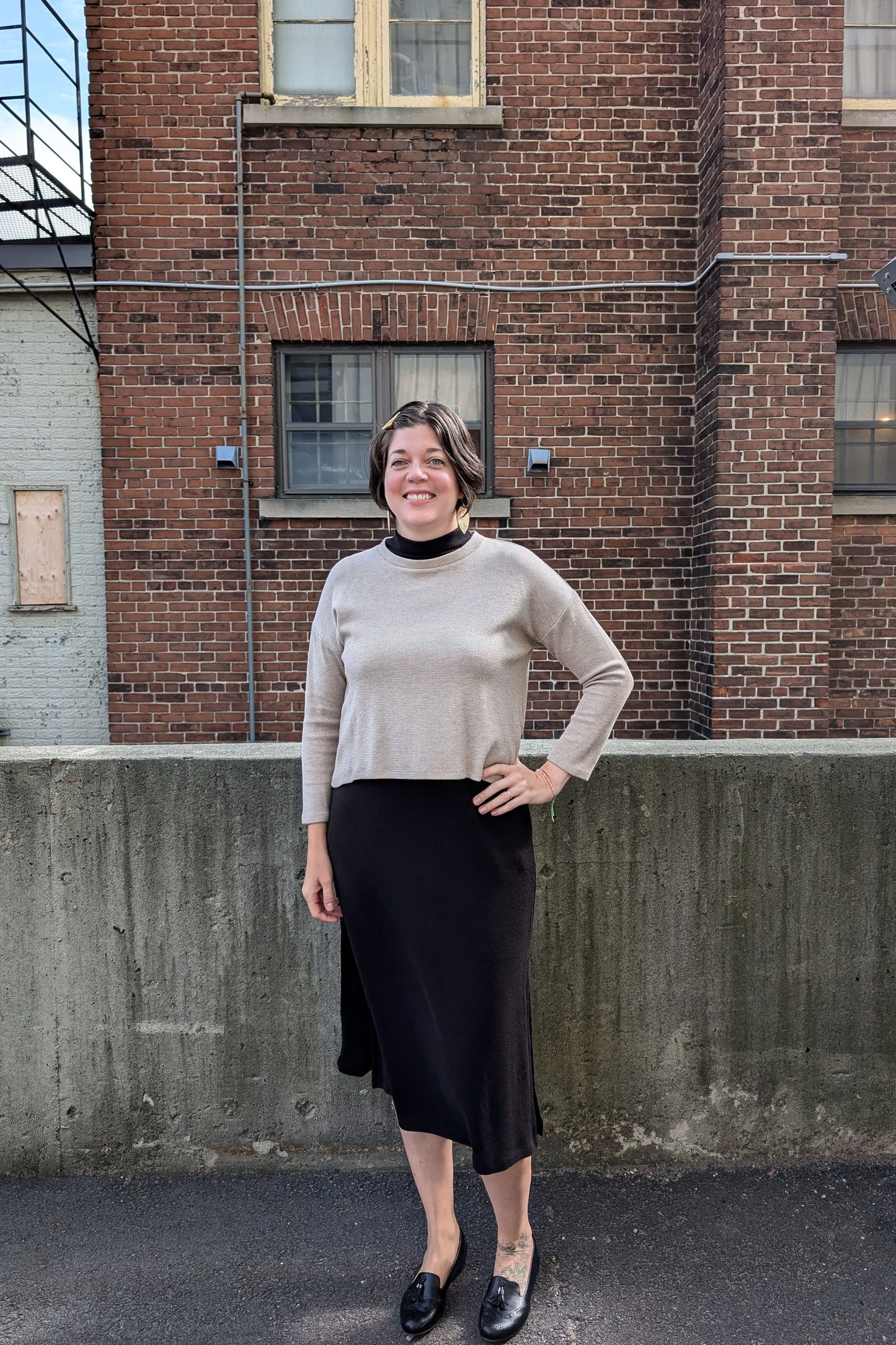 A woman wearing the Nerissa Knit Top by Compli K in Beige, with a round neck, long sleeves, slightly loose fit, and hi-low hemline, over the Natasha Dress in Black. She is standing outside in front of a brick wall.
