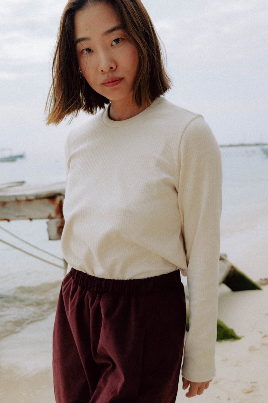 A woman wearing the Whistler Long Sleeve Top in Almond, a ribbed cotton knit with a round neck and straight cut. She is wearing it with the Funchal Pants in Fig and standing on a beach.