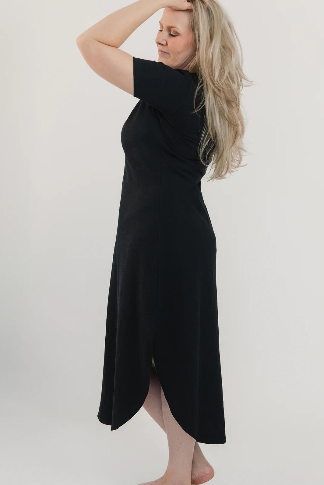 Side view of a woman wearing the Field Dress by Blondie in Black, with short sleeves, a scooped neck, and a contoured hem that hits at mid-calf and cuts up at the sides. She is standing in front of a white background. 