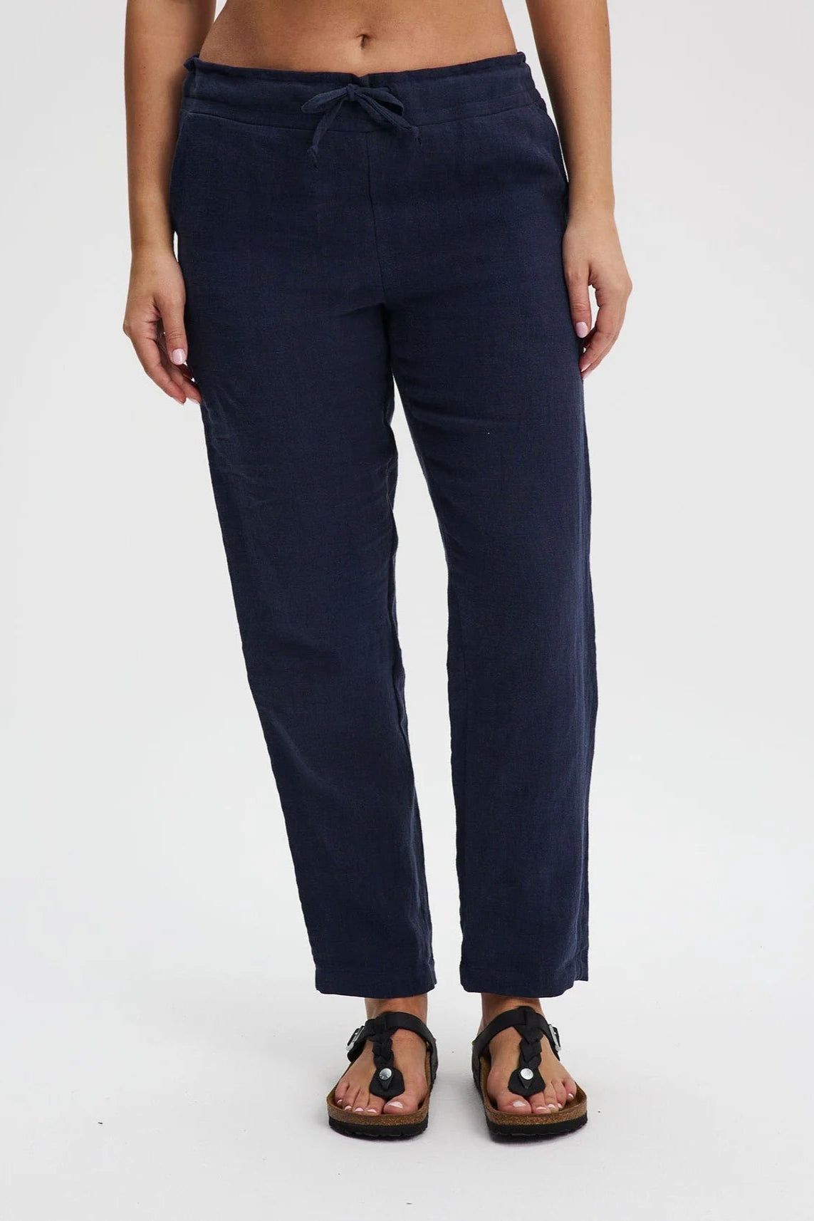 A waist down view of a woman wearing the Providence Pants by Message Factory in Navy, 100% linen pants with a slim fit, drawstring waist, and front and back pockets. She is standing in front of a white background. 