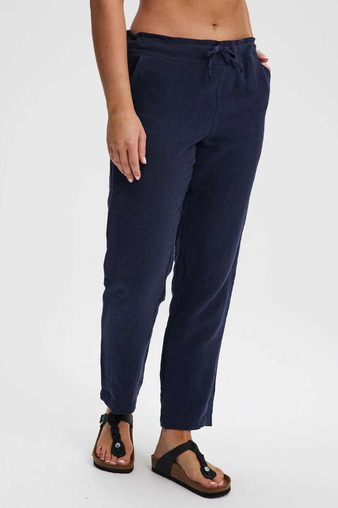 A waist down view of a woman wearing the Providence Pants by Message Factory in Navy, 100% linen pants with a slim fit, drawstring waist, and front and back pockets. She is standing in front of a white background. 