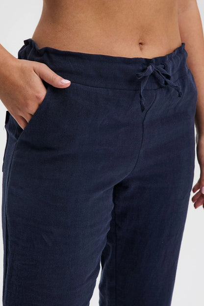 A waist down close-up of a woman wearing the Providence Pants by Message Factory in Navy, 100% linen pants with a slim fit, drawstring waist, and front and back pockets. She is standing in front of a white background. 