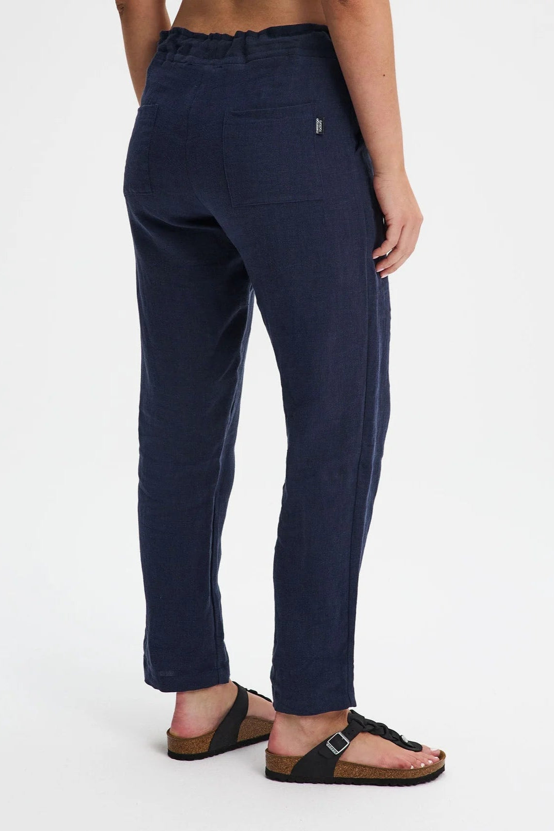 A waist down back view of a woman wearing the Providence Pants by Message Factory in Navy, 100% linen pants with a slim fit, drawstring waist, and front and back pockets. She is standing in front of a white background. 