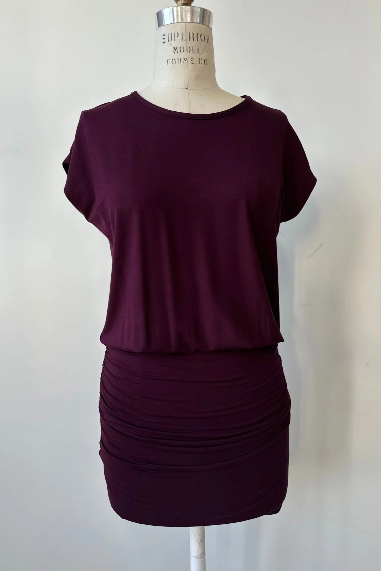 The Gabriel Tunic by Cherry Bobin in Burgundy is shown on a mannequin in front of a white background. It features gathers at the lower sides for an adjustable length, a wide neck, and short extended sleeves. 