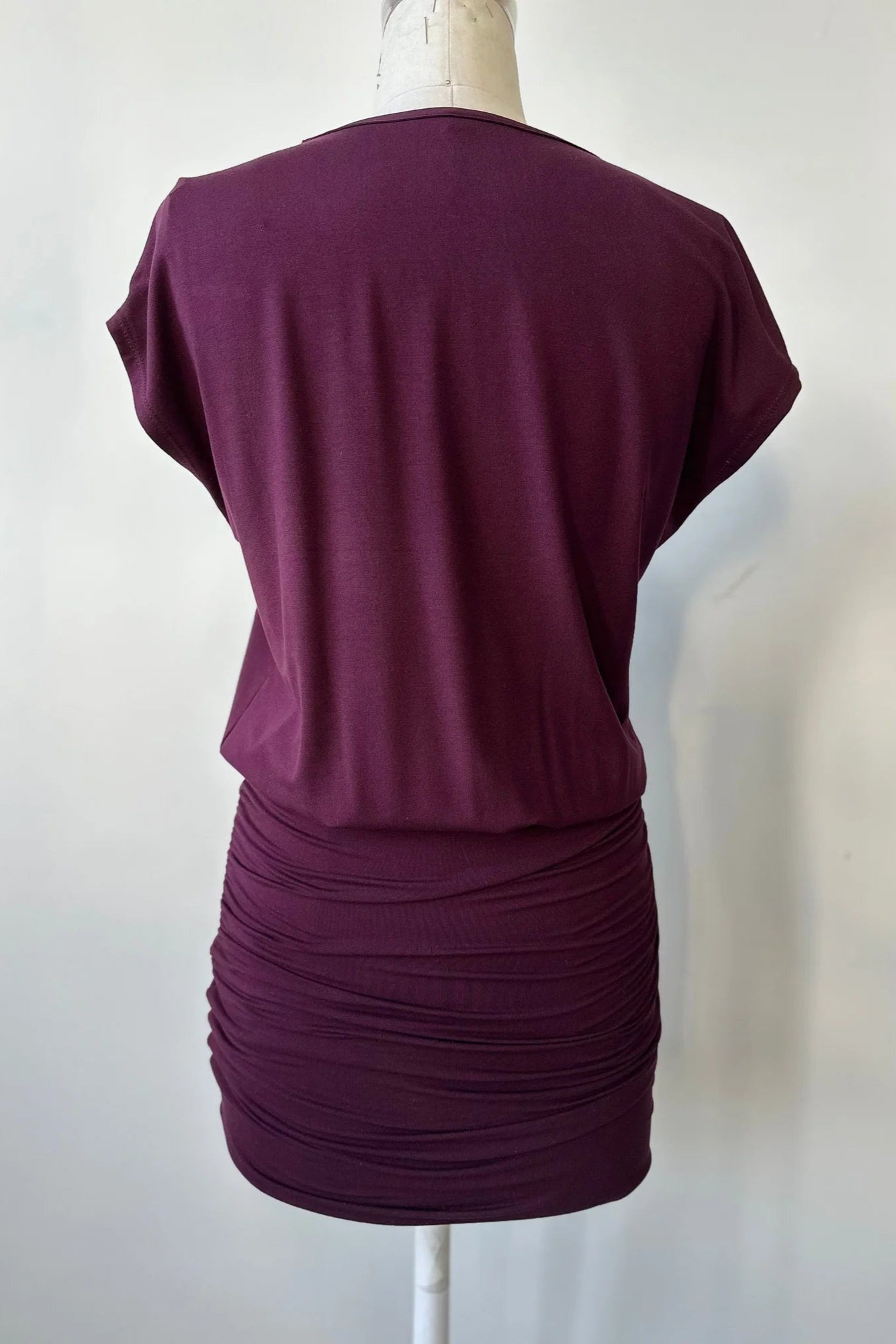 A back view of the Gabriel Tunic by Cherry Bobin in Burgundy is shown on a mannequin in front of a white background. It features gathers at the lower sides for an adjustable length, a wide neck, and short extended sleeves. 