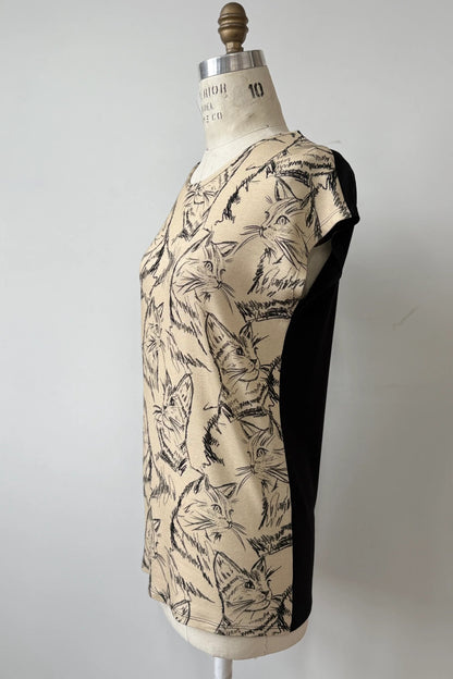 A side view of the Arto Tunic by Kollontain in Cats/Beige Print is shown on a mannequin against a grey background. It has a round neck, short extended sleeves, and a straight cut with a solid black back.  