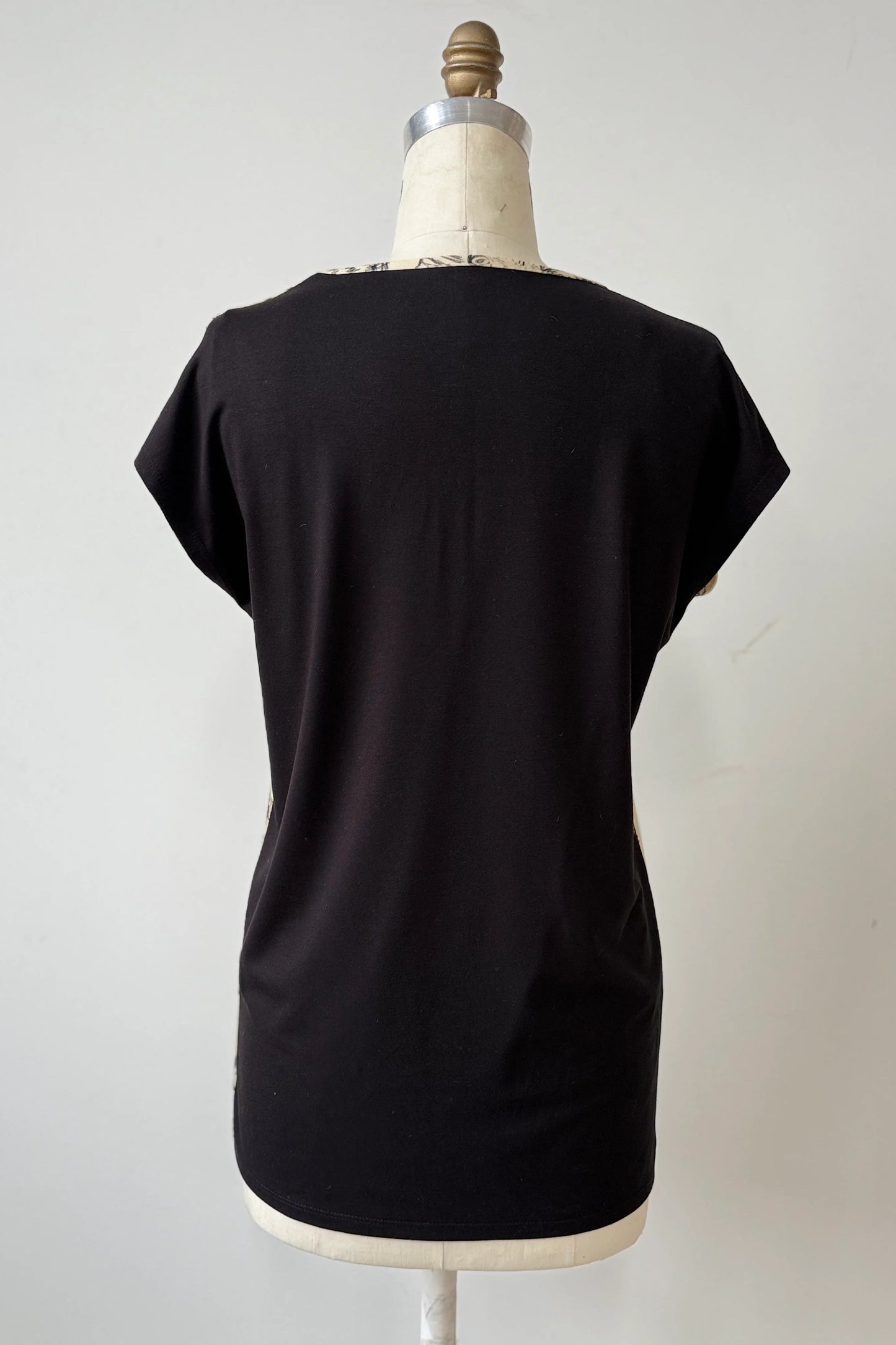 A back view of the Arto Tunic by Kollontain in Cats/Beige Print is shown on a mannequin against a grey background. It has a round neck, short extended sleeves, and a straight cut with a solid black back. 