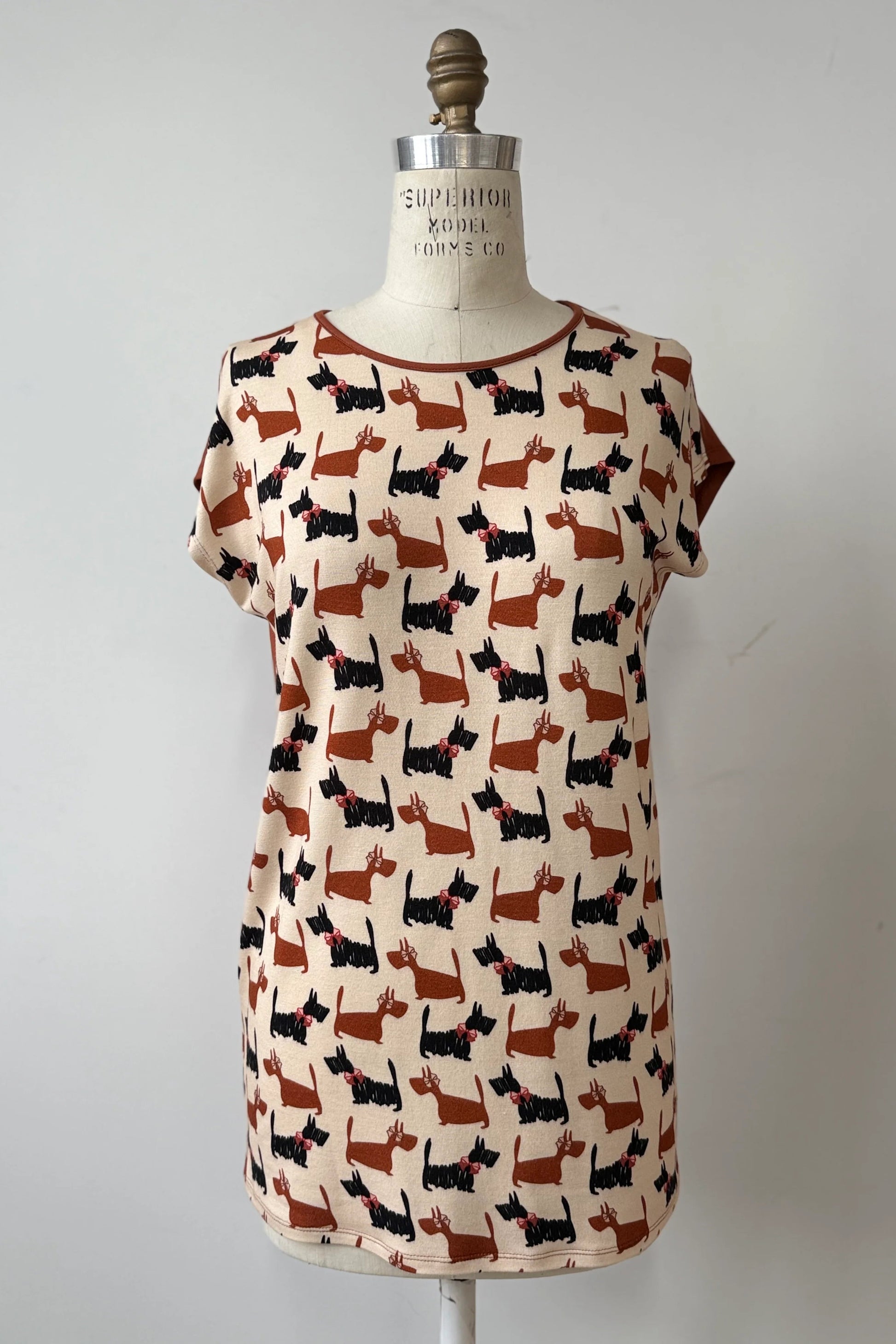 The Arto Tunic by Kollontai in Dogs/Rust Print is shown on a mannequin against a grey background. It has a round neck, short extended sleeves, and a straight cut with a solid rust coloured back. 