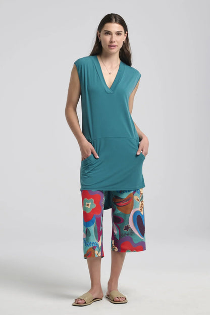A woman wearing the Morticia Dress by Kollontai in Ocean, a sleeveless dress with a V-neck, a hi-low hemline, and a front pouch pocket. She is wearing it with patterned cropped pants standing in front of a white background. 