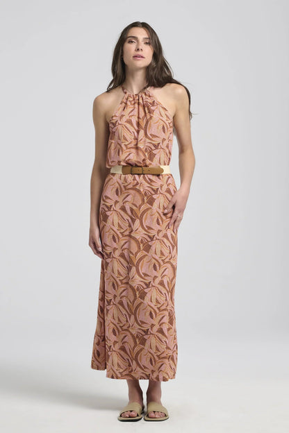 A woman wearing the Anwen Dress by Kollontai in Cinnamon print, a loose-fitting viscose maxi dress with an adjustable halter neckline. She is wearing it with a belt and is standing in front of a white background. 