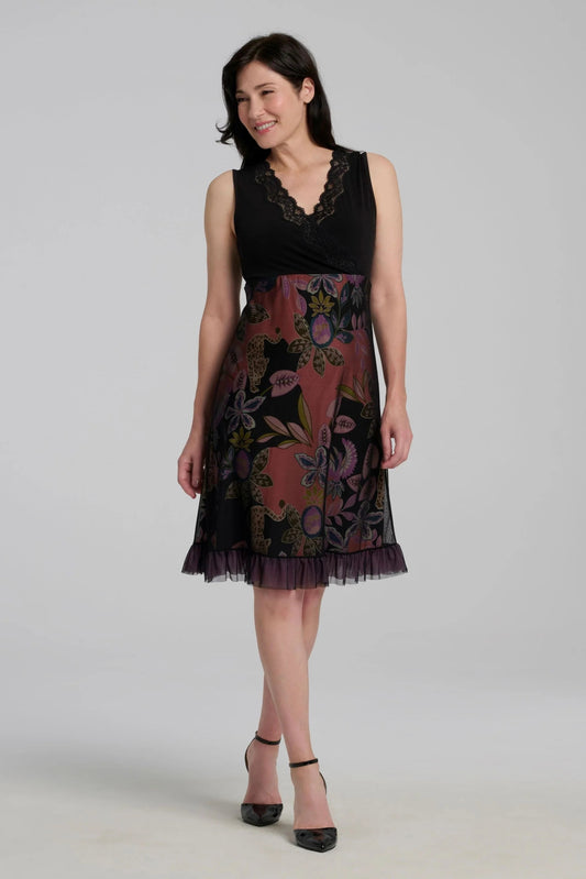 A woman wearing the Liseron Dress by Kollontai, a sleeveless dress with a faux-wrap neckline, empire waist, knee-length a-line skirt. The bodice is solid black, while the skirt features a cheetah/floral print under a mesh overlay. She is standing in front of a grey background. 