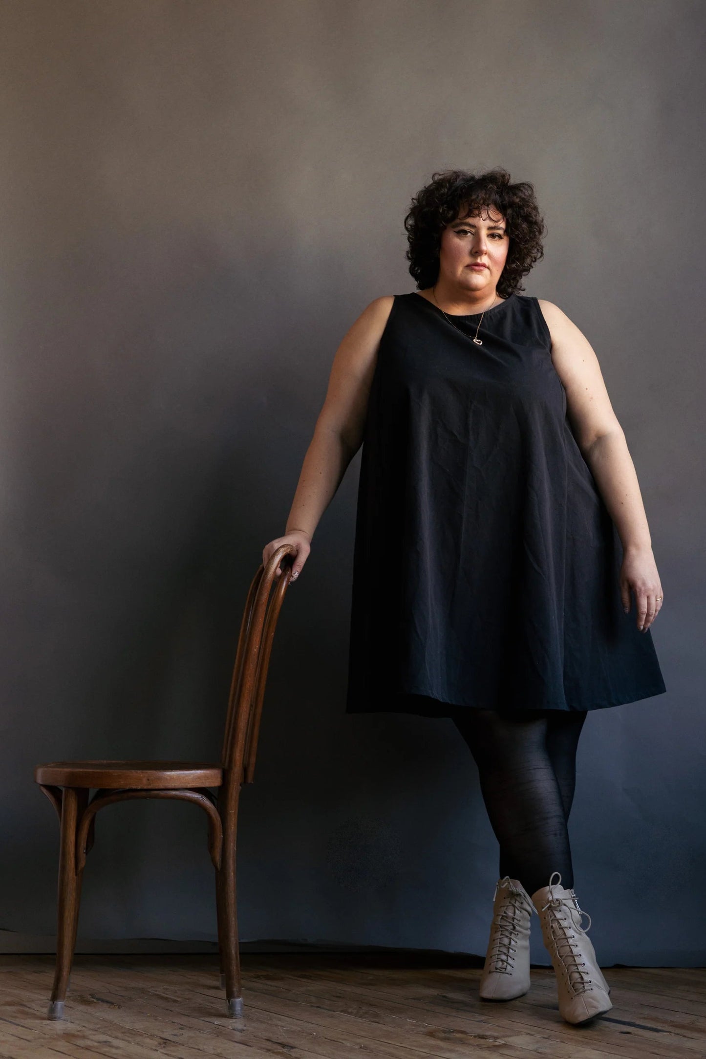 A woman wearing the Caro Dress by Lights of All in Black, a sleeveless knee-length trapeze dress with a round neck at the front and a V at the back. She is wearing it with black leggings and standing with her arm resting on a chair in front of a dark background. 