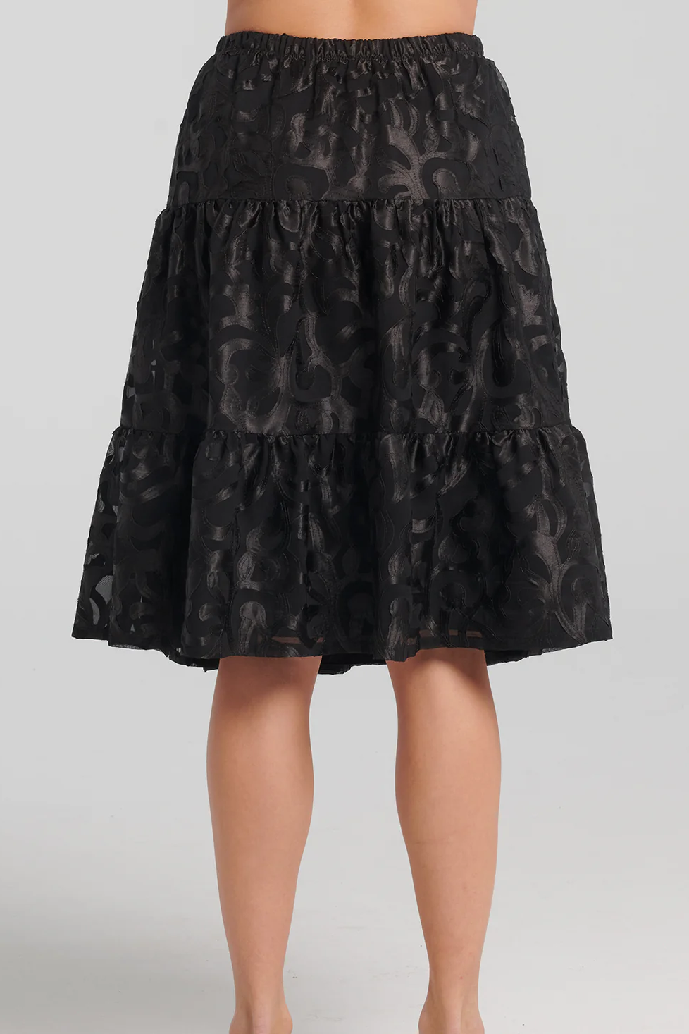 Back waist-down view of a woman wearing the Zimmer Skirt by Kollontai in Black, standing in front of a grey background