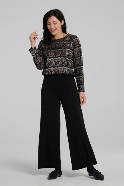 A woman wearing the Solstice Blouse by Kollontai in Gold, with fabric that layers black lace over golden satin. It's slightly cropped with an elastic waist and long sleeves. She is wearing it with black pants and standing in front of a grey background. 