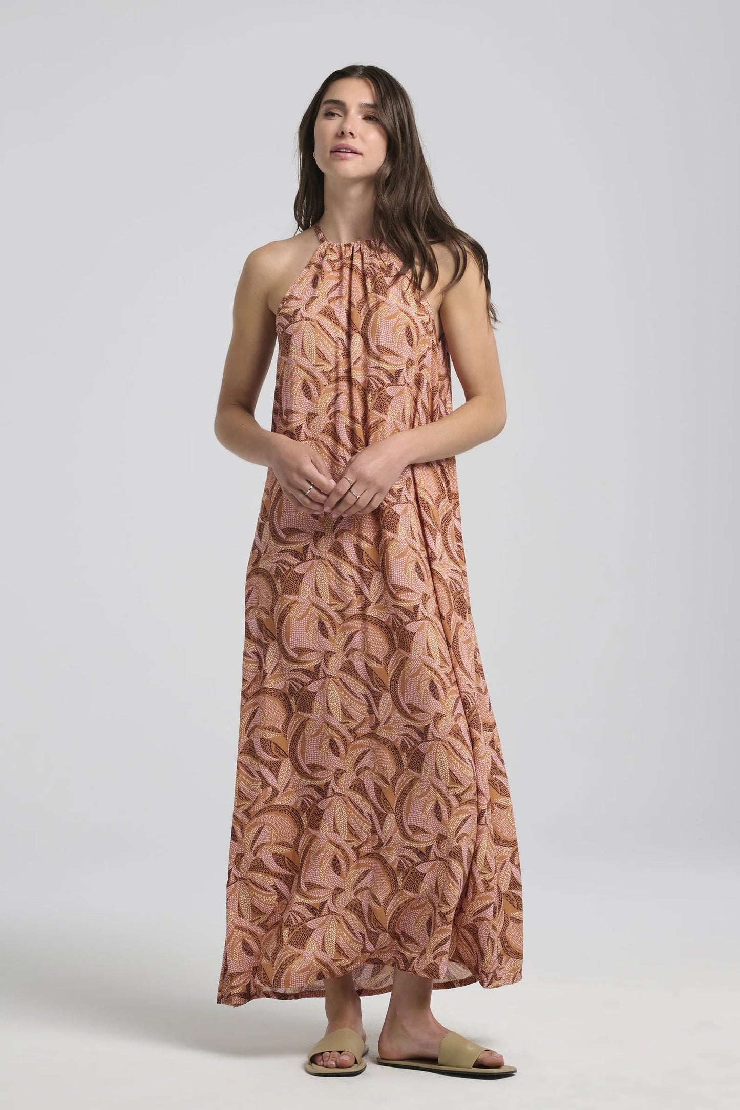 A woman wearing the Anwen Dress by Kollontai in Cinnamon print, a loose-fitting viscose maxi dress with an adjustable halter neckline. She is standing in front of a white background. 