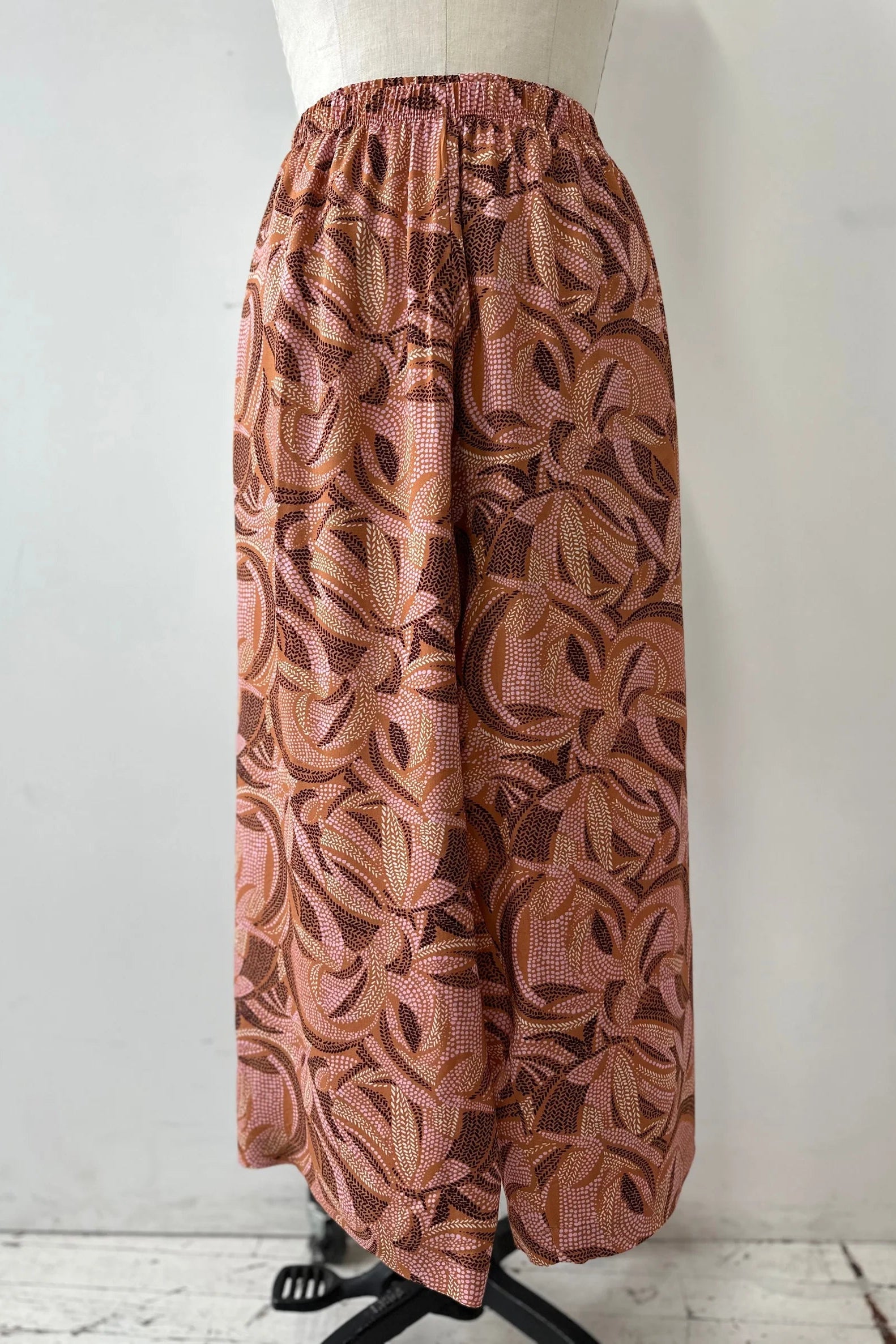 The Isiclora Pants by Kollontai in Cinnamon print, a lightweight pant with an elastic waist and flowing, cropped legs, are show on a mannequin in front of a white background. 