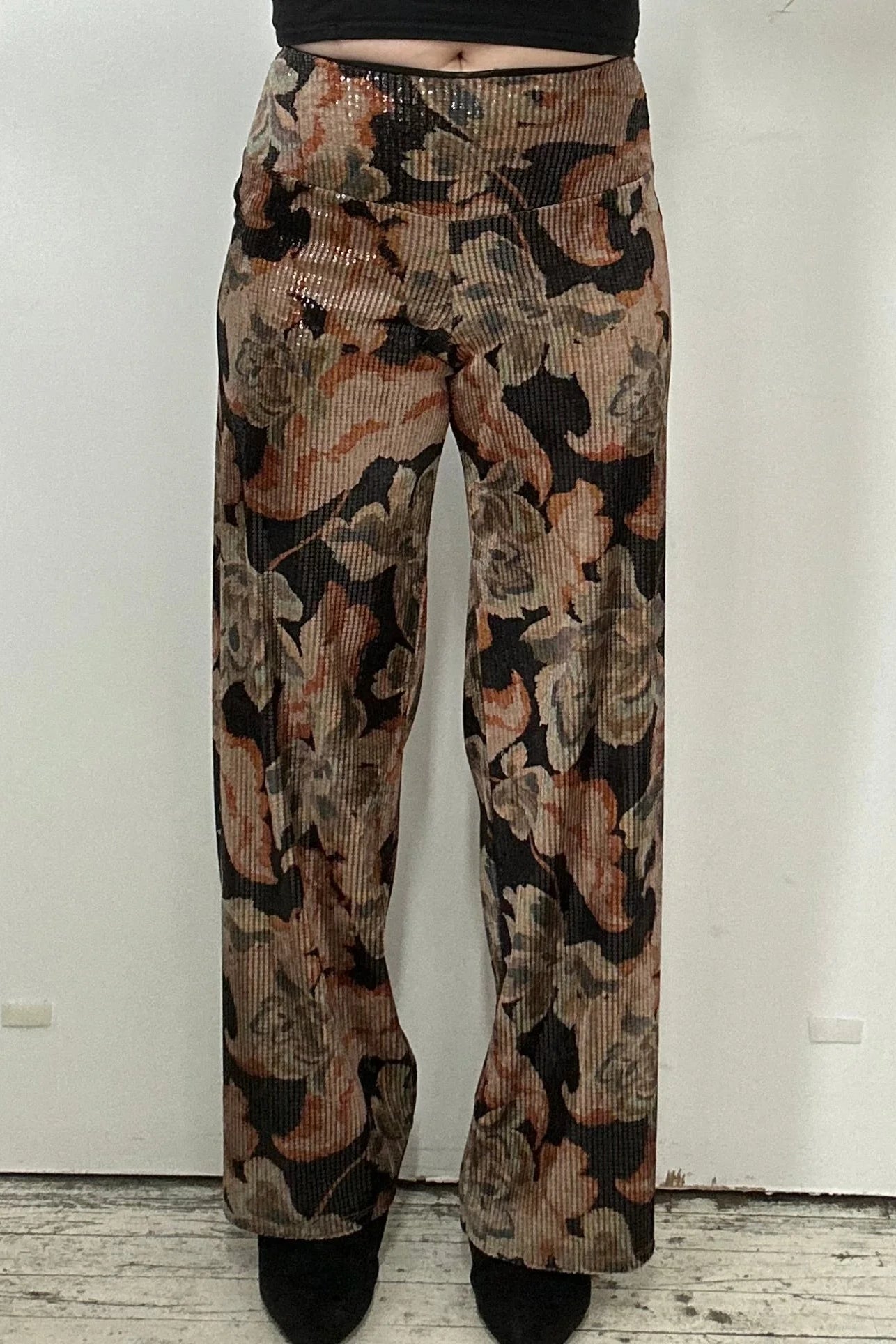 Waist down view of a woman wearing the Carina Pants by Kollontai, featuring floral velvet in autumn tones with clear sequins sewn right into the fabric, full-length straight legs and a wide pull-on waistband. She's wearing them with a black top and standing in front of a white background. 
