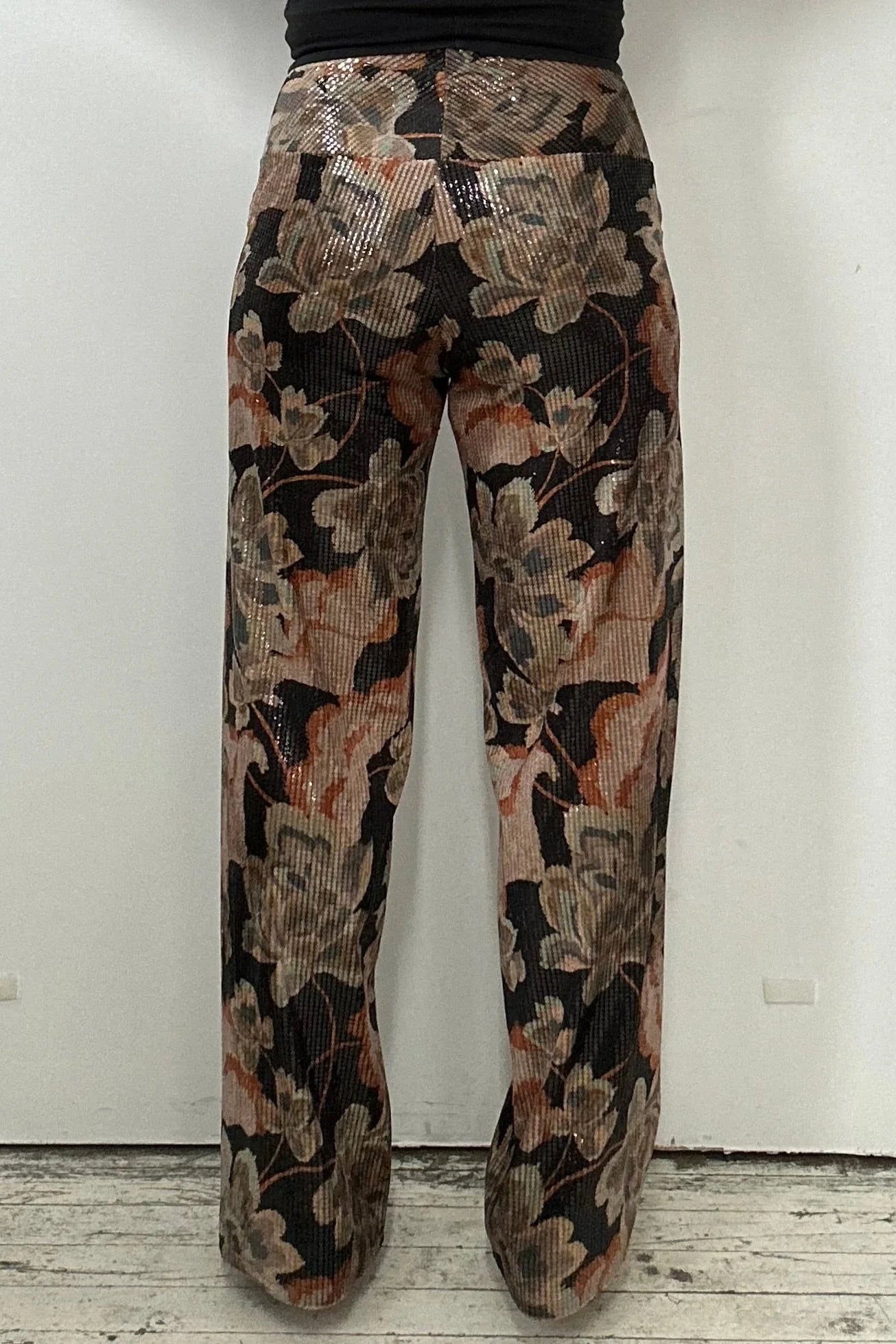 Back waist down view of a woman wearing the Carina Pants by Kollontai, featuring floral velvet in autumn tones with clear sequins sewn right into the fabric, full-length straight legs and a wide pull-on waistband. She's wearing them with a black top and standing in front of a white background. 