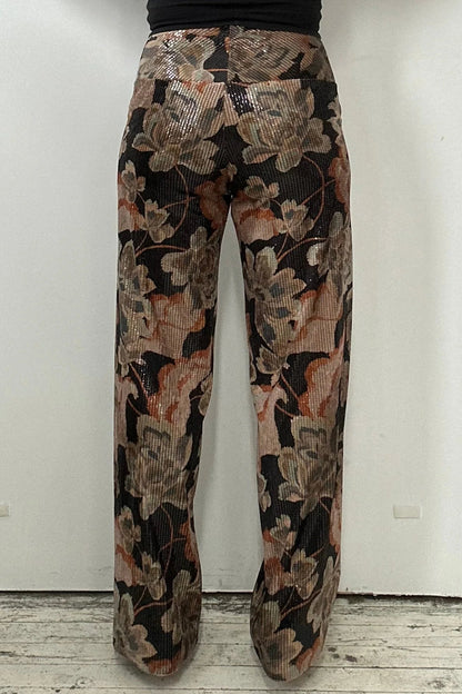 Back waist down view of a woman wearing the Carina Pants by Kollontai, featuring floral velvet in autumn tones with clear sequins sewn right into the fabric, full-length straight legs and a wide pull-on waistband. She's wearing them with a black top and standing in front of a white background. 